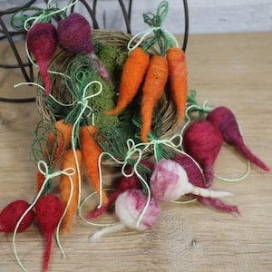 May include: A collection of felt carrots and beets, tied with twine and green stems. The vegetables are arranged in a small woven basket with moss and are displayed on a wooden surface.