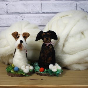 May include: Two needle felted dog figurines, one white and brown, the other black and brown, sitting on a green base with colorful Easter eggs. The dogs are surrounded by white wool roving.