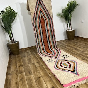 Op de afbeelding: Een crèmekleurige, rechthoekige loper met een centraal geometrisch ontwerp in tinten oranje, roze, teal en zwart. Het tapijt heeft een roze franje en ligt op een houten vloer.