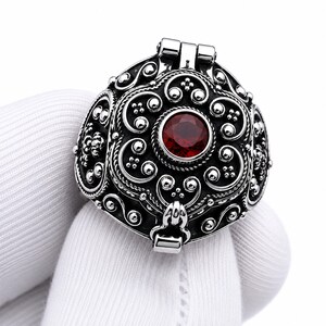 May include: A circular silver locket ring featuring an intricate oxidized metal filigree pattern with swirling scrolls and small spheres. A single round, faceted deep red gemstone is set in the center of the hinged top cover of the ring.