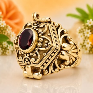Könnte beinhalten: Ein goldfarbener Ring mit einem ovalen, dunkelroten Edelstein in der Mitte. Die Ringschiene und die Fassung sind mit detaillierten, erhabenen Schnörkeln, Filigranmustern und kleinen Perlenakzenten verziert, wobei ein Scharniermechanismus sichtbar ist.