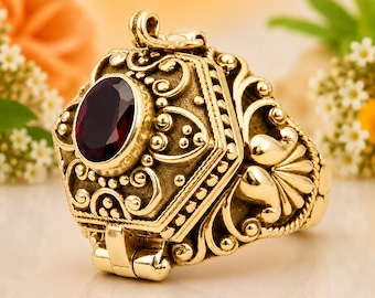 May include: A gold-toned ring featuring an oval-shaped dark red gemstone set in the center. The band and setting are decorated with intricate raised scrollwork, filigree patterns, and small bead accents, with a hinged mechanism visible.