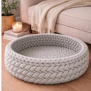 May include: A light grey, round pet bed with a braided design. The bed is made of a soft, chunky knit material. The bed is on a hardwood floor.