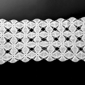 Può includere: Runner da tavolo in pizzo bianco a trama aperta con motivo floreale ripetuto. Il runner è rettangolare con un bordo smerlato. Il design intricato è visibile su uno sfondo nero.