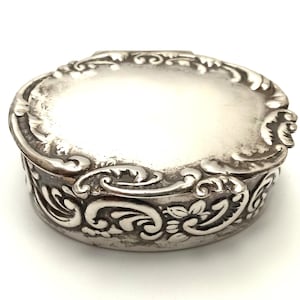 May include: A small, oval-shaped silver trinket box featuring an ornate, raised floral and scrollwork pattern along the lid's edge and the sides. The metal surface has a polished finish with darkened recessed areas highlighting the intricate details.