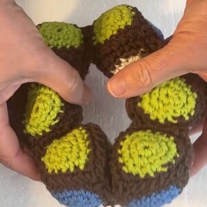 Könnte beinhalten: Ein handgefertigter Häkelkranz mit einem sich wiederholenden Muster aus braunem, grünem und blauem Garn. Der Kranz wird von zwei Händen gehalten, die die Textur und Farbvariationen des Garns hervorheben. Dieser Dekorationsartikel eignet sich für die Inneneinrichtung.