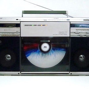 Może przedstawiać: Srebrno-czarny, zabytkowy boombox z odtwarzaczem kaset, radiem i odtwarzaczem CD. Boombox ma dwa duże głośniki i wysuwaną antenę. Odtwarzacz CD jest widoczny pośrodku, z kolorowym wzorem.