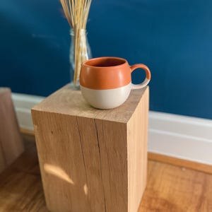 May include: A two-tone ceramic mug with a rounded shape, featuring a burnt orange top and a cream-colored bottom. The mug sits on a natural wood block, with dried flowers in a glass vase in the background.