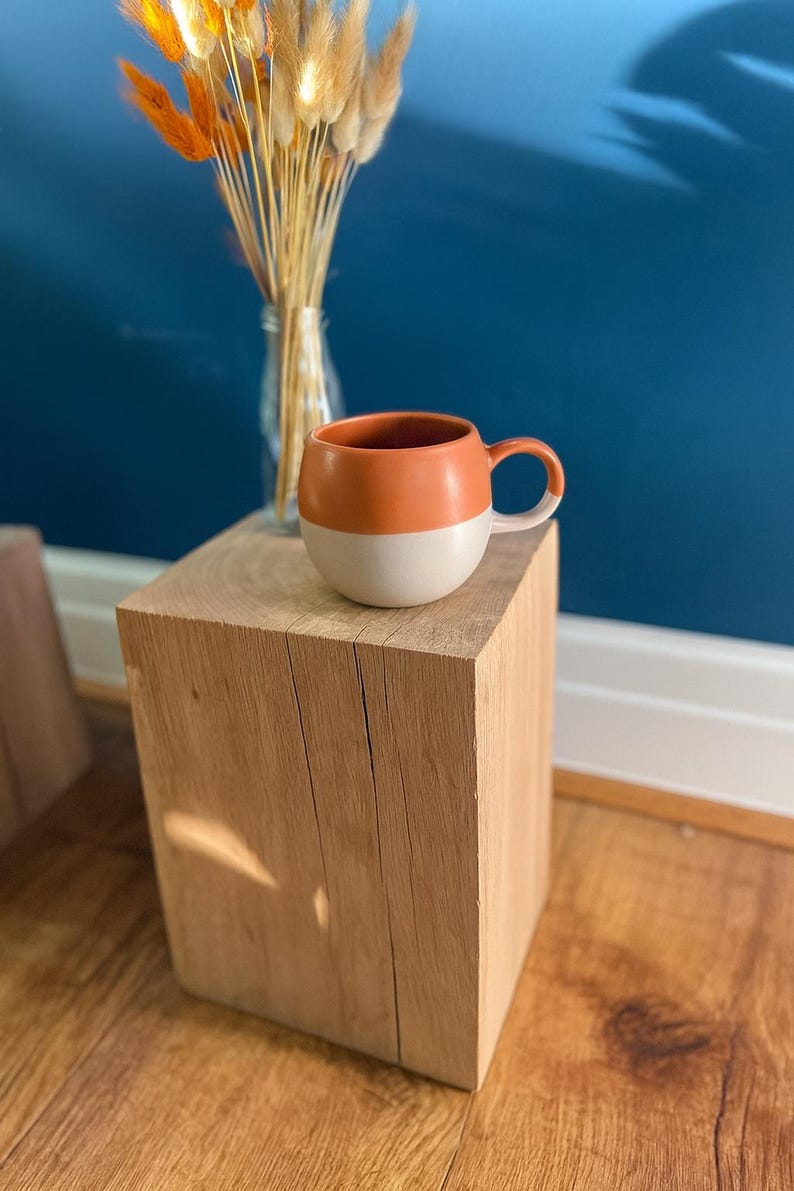 May include: A two-tone ceramic mug with a rounded shape, featuring a burnt orange top and a cream-colored bottom. The mug sits on a natural wood block, with dried flowers in a glass vase in the background.