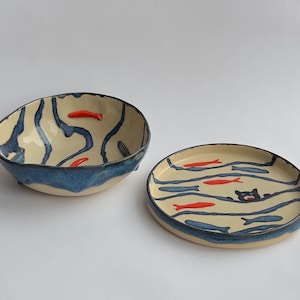 Op de afbeelding: Een keramische kom en bord set. De kom heeft een crèmekleurig interieur met blauwe golfachtige patronen en rode visaccenten. Het bord heeft een vergelijkbaar ontwerp, met een zwarte kat die tussen de vissen zwemt. Beide stukken hebben blauwe randen.