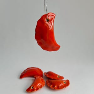 Pode incluir: Um conjunto de ornamentos de pierogi de cerâmica vermelha. Um pierogi está suspenso por uma corrente prateada. Os outros quatro pierogi estão sobre uma superfície branca. Cada pierogi tem um acabamento brilhante e um pequeno orifício.