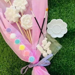 May include: A craft kit presented as a bouquet, with white flower and butterfly-shaped items on sticks, paint pots in various colours, paintbrushes, and a palette. The bouquet is wrapped in pink tissue paper and tied with a purple and white ribbon.