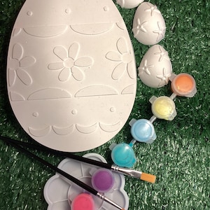 May include: A large, unpainted Easter egg and several smaller eggs with flower designs. Small pots of paint in various colours, two paintbrushes, and a flower-shaped paint palette are also included. The items are arranged on a green surface.