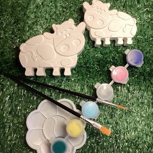 May include: Two white cow-shaped figurines with black spots, ready to be painted. Includes a paint palette with various colors, two paintbrushes, and paint pots. The set is designed for creative painting projects.