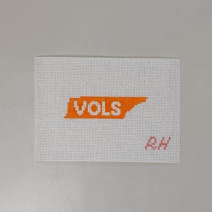 May include: A white needlepoint canvas featuring an orange silhouette of the state of Tennessee with the white text VOLS centered inside. The letters RH are stitched in light pink in the bottom right corner of the rectangular grid canvas.