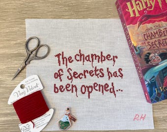 May include: A square piece of white needlepoint canvas featuring the dark red stitched text The chamber of Secrets has been opened, accompanied by a pair of vintage-style scissors, a spool of red Very Velvet thread, and a small potion bottle pin.