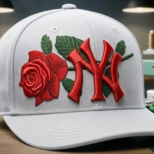 May include: A white baseball cap featuring a red rose and the letters "NY" in red. The rose and letters are embroidered. The cap is on a wooden surface, with spools of thread and an embroidery machine in the background.