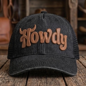 Puede incluir: Una gorra de camionero de mezclilla negra con parte trasera de malla y panel frontal estructurado. La palabra Howdy aparece en letras marrones en relieve, similares al cuero, en el centro del panel frontal, con la H en forma de cuernos.