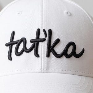 Puede incluir: Gorra de béisbol blanca con la palabra "tat'ka" bordada en negro en la parte delantera. La gorra tiene una visera curva y una corona estructurada. Otra gorra blanca es visible en el fondo.