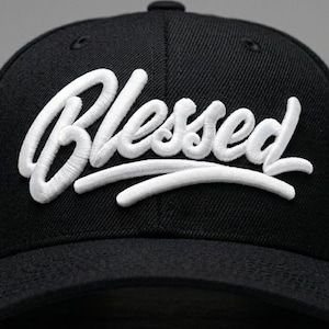 Pode incluir: Boné de beisebol preto com a palavra "Blessed" bordada em branco na frente. O texto está em escrita cursiva. O boné tem uma viseira curva.