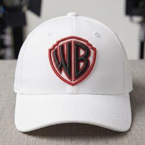 Puede incluir: Gorra de béisbol blanca con el logotipo de Warner Bros. El logotipo tiene forma de escudo con borde rojo y letras negras "WB". La gorra tiene una visera curva y está hecha de tela blanca lisa.