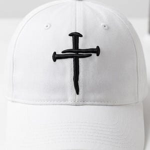 May include: White baseball cap with a black embroidered cross made of nails. The cap has a curved brim and a structured crown. The design is centered on the front of the cap.