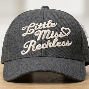 May include: A charcoal gray baseball cap with the embroidered text "Little Miss Reckless" in white script. The cap has a curved brim and a small button on top. A white tag is attached to the side.
