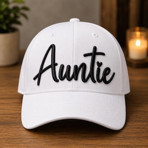 May include: A white baseball cap featuring the word Auntie in raised black lettering across the front. The text is written in a cursive script font, and a small heart icon is integrated into the design above the letter i in the word Auntie.