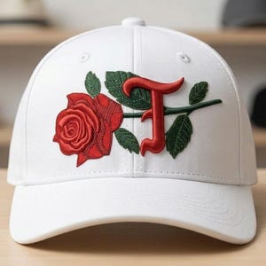 Puede incluir: Gorra de béisbol blanca con una rosa roja y la letra "J" bordada. La rosa y la letra son rojas, con hojas y tallo verdes. La gorra tiene una visera curva. El diseño es floral y con inicial.