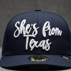 May include: A navy blue baseball cap with the embroidered text "She's From Texas" in white script. The cap has a curved brim and a structured crown. The hat is a 59FIFTY style.