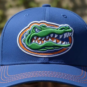 May include: A blue baseball cap featuring an embroidered alligator logo in green and orange. The cap has a curved peak and orange stitching. A classic style hat, suitable for sports fans.