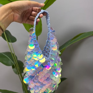 May include: A small, pale blue crocheted handbag decorated with iridescent sequins. The sequins reflect a spectrum of colours, creating a shimmering effect. The bag has a woven handle and is set against a backdrop of green foliage.