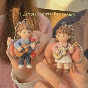 Puede incluir: Dos llaveros con figuras en miniatura de un niño y una niña tocando la guitarra. El niño lleva una camisa azul y pantalones oscuros, mientras que la niña viste un top rosa y una falda blanca. El texto "CUSTOM COUPLE KEYCHAINS!" es visible.