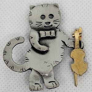 Peut inclure: Broche en forme de chat argenté avec un nœud papillon, tenant un violon doré. Le chat a des détails gravés et un visage souriant. Le violon est détaillé avec des cordes et un archet.