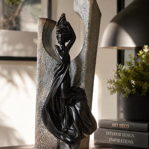 May include: A tall sculpture featuring a black figure with flowing fabric draped around the body, positioned in front of a textured, weathered gray and bronze-toned archway. The entire piece is mounted on a solid black rectangular base.