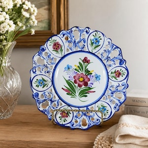 Peut inclure: Assiette décorative en céramique blanche avec un motif floral bleu et une bordure ajourée. L'assiette présente un arrangement floral central dans les tons de rouge, violet et bleu. Elle est présentée sur un support doré.