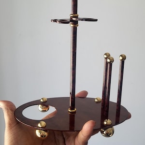 May include: A decorative, multi-tiered stand with a brown, wood-grain finish and gold accents. The stand features a circular base, vertical supports, and a gold sphere at the top. The stand is held in a hand.