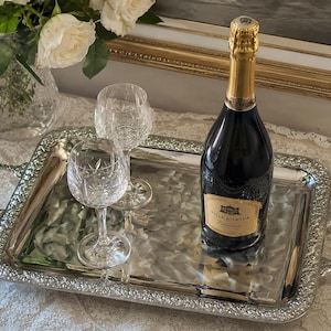 Può includere: Un vassoio da portata rettangolare in metallo argentato con manici e bordi goffrati, contenente una bottiglia di vetro scuro di Villa Jolanda Prosecco con collo dorato e due calici in cristallo trasparente con motivo intagliato.