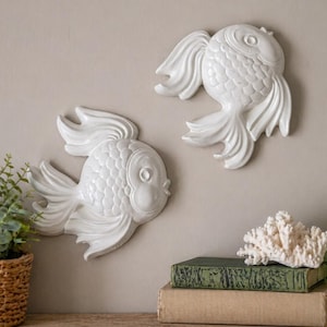 May include: Two white, decorative fish wall hangings with detailed scales and flowing fins. The fish are mounted on a neutral wall. A wicker basket with greenery, books, a coral piece, and a green glass bottle are on a shelf below.
