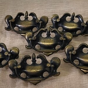 May include: Six antique brass drawer pulls with an ornate design. Each pull has a curved handle, central finial, and rounded accents. The pulls are arranged on a neutral fabric background. Ideal for furniture restoration.