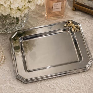 May include: A rectangular silver-toned metal tray with beveled edges and a polished reflective surface. A small, textured gold-toned metal bow ornament is attached to the top right corner of the tray, adding a decorative detail to the metallic frame.