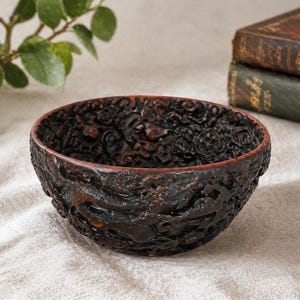 May include: A dark brown, intricately carved bowl with a textured surface. The bowl's exterior features detailed relief patterns, and the interior has a reddish-brown hue. The bowl is set against a neutral background.