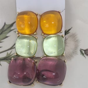 May include: A pair of square drop earrings with gold-colored settings. Each earring features three stacked, square-shaped stones in shades of amber, light green, and purple. The earrings are displayed on a white background.