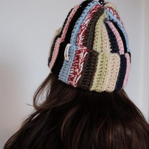 Può includere: Un berretto a righe multicolore nei toni del rosa, nero, blu, rosso, marrone e beige. Il cappello fatto a mano è indossato sulla testa di una persona. Una piccola etichetta con la scritta "elsefrida" è attaccata.