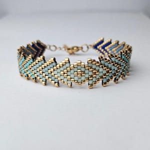Peut inclure: Un bracelet de perles avec un motif en chevrons. Le bracelet présente des perles dorées et des rangées alternées de perles bleu clair et bleu foncé. Le bracelet mesure environ 18 cm de long.