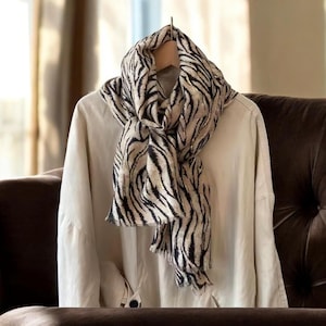 May include: A beige and black zebra print scarf draped over a light-colored shirt on a wooden hanger. The scarf's pattern features bold black stripes against a beige background. The shirt is a neutral color, creating a contrast with the scarf.