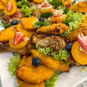 May include: A platter of golden-brown breaded chicken cutlets and roasted chicken pieces garnished with fresh green lettuce, vibrant yellow peach halves, sliced strawberries, and dark blue blueberries, accented with scattered pomegranate seeds.