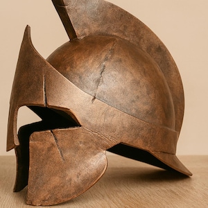 Peut inclure: Casque spartiate de couleur bronze, avec une crête et des protège-joues distinctifs. Le casque présente un aspect vieilli, avec une texture et des lignes visibles, suggérant une esthétique ancienne. Le design rappelle les casques militaires grecs antiques.