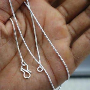 May include: A thin, silver-coloured snake chain necklace featuring a smooth, flexible metallic texture. The necklace includes a simple S-hook clasp mechanism at one end and a small circular jump ring at the other end for secure fastening.