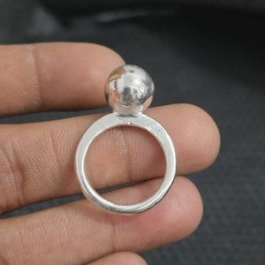 Pode incluir: Um anel de prata com uma esfera polida sobre uma banda circular. O anel é segurado em uma mão, mostrando seu design simples. A superfície refletora do anel capta a luz, destacando sua textura lisa.